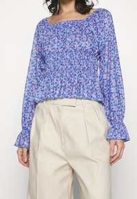 Bluse mit blauem Blumenmuster, gerafftem Oberteil und langen Ärmeln, mit gerafften Manschetten. Getragen mit hoch taillierten beigen Hosen.
