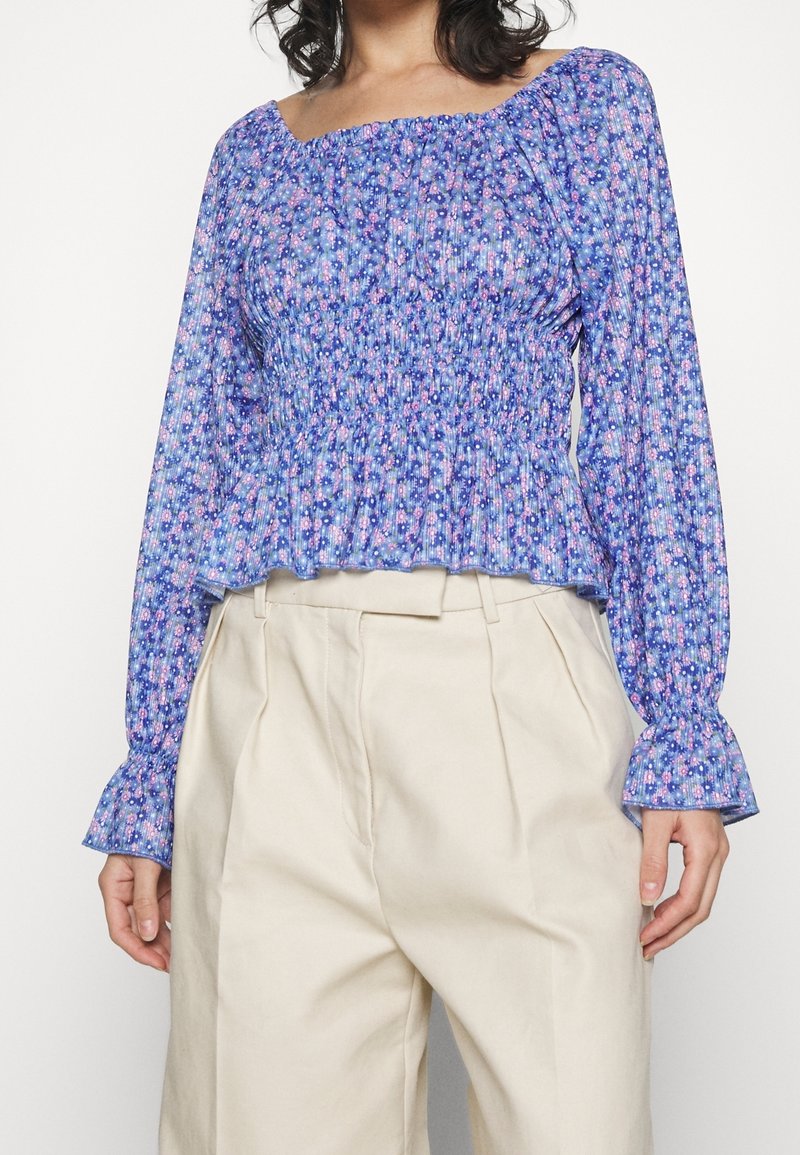 Bluse mit blauem Blumenmuster, gerafftem Oberteil und langen Ärmeln, mit gerafften Manschetten. Getragen mit hoch taillierten beigen Hosen.