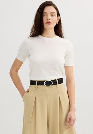 Mujer con cabello largo y castaño que viste una camiseta blanca de manga corta, pantalones beige plisados y un cinturón negro con hebilla plateada.