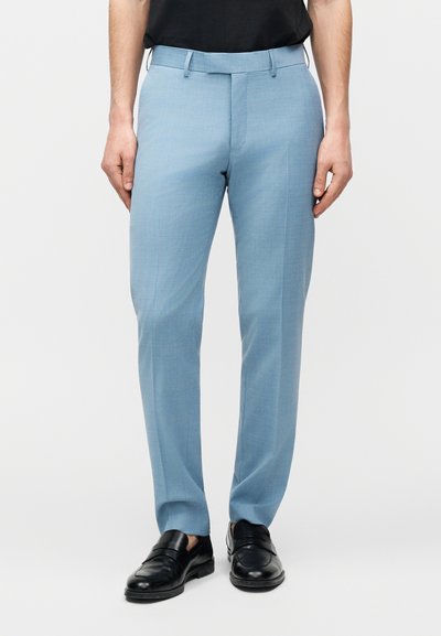 Άνδρας φοράει ανοιχτό μπλε παντελόνι ραμμένο στα μέτρα και μαύρα δερμάτινα μοκασίνια, στέκεται μπροστά σε ένα απλό φωτεινό φόντο.