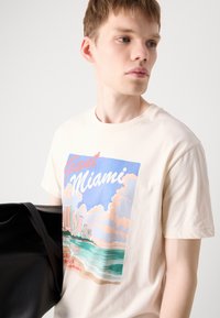 Heledadbež värvi puuvillane T-särk, millel on graafiline print Miami siluetist ja pilvedest pastellvärvides, ümar kaelus ja lühikesed varrukad.