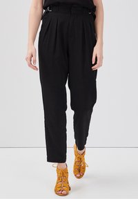 Person wearing high-waisted black pleated pants and mustard yellow lace-up open-toe heels on a light background.