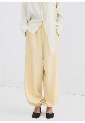 Mujer con pantalones sueltos de color crema con puños elásticos y zapatos negros de tacón, de pie con las manos en los bolsillos sobre un fondo blanco.