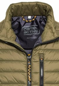 Olivgrüne Steppjacke mit steigendem Kragen, schwarzer Innenseite und Frontreißverschluss. Enthält recycelte Materialien und ein Logo innen.