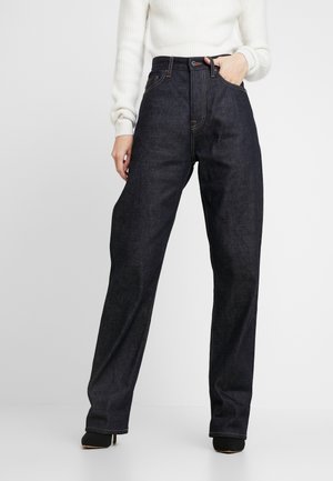 Woman wearing high-waist dark blue wide-leg jeans and a white ribbed sweater, one hand in pocket, standing against a white background.