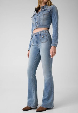Femme portant une veste en jean courte bleu clair et un jean évasé taille haute avec des chaussures marron pointues sur fond uni.