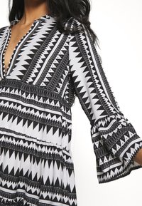 Svart och vit mönstrad klänning med V-ringning, utsvängda ärmar och geometriska mönster, tillverkad av mjukt tyg.