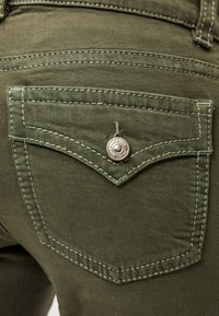 Πράσινο τζιν ύφασμα με πίσω τσέπη, που διαθέτει ασημένιο κουμπί και αντίθετη ραφή. Η τσέπη έχει καμπυλωτό, γωνιακό σχέδιο.