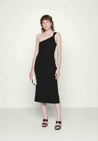 Vestido midi preto com um ombro, de silhueta ajustada e fenda lateral. Modelo segura uma clutch texturizada branca e usa sandálias pretas.