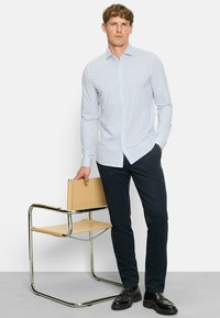 Hellblaues Hemd mit einem kleinen geometrischen Muster, kombiniert mit dunklen Hosen und schwarzen Schuhen. Das Model steht neben einem minimalistischen Stuhl.
