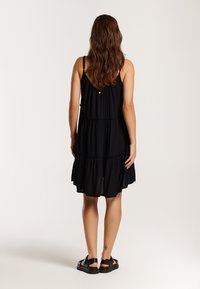 Femme aux longs cheveux bruns portant une robe noire sans manches à volants et des sandales noires, debout de dos sur un fond uni.