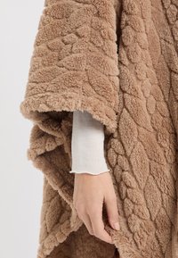 Przytulny beżowy ponczo z polaru o fakturowanym wzorze. Miękki i puszysty materiał opada na białą koszulę z długim rękawem, z falistym brzegiem.