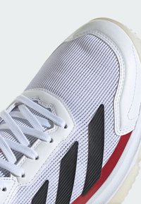 Baskets blanches de sport avec une tige en maille texturée, des accents noirs et rouges, et une semelle rembourrée. Les lacets et les superpositions en matériau synthétique complètent le design.