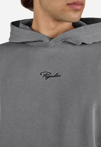 Young person wearing a gray hoodie with a small black embroidered "Pegador" logo on the chest, facing slightly right.