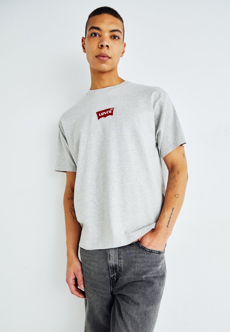 Szürke pamut póló rövid ujjakkal, a mellkason piros Levi's logóval. Sötétszürke farmernadrággal párosítva.