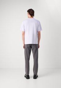 Mężczyzna stoi tyłem, ubrany w jednolitą jasnopurpurową koszulę z krótkim rękawem, szare dżinsy i czarne buty na jednolitym tle.