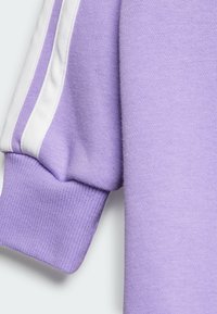 Lila pulóver ujja fehér díszcsíkokkal, bordázott mandzsettával és struktúrált anyaggal, amely sima, puha megjelenést kölcsönöz.