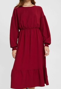 Robe rouge à manches longues avec une coupe ample, taille froncée et ourlet volé. Tissu texturé avec un aspect doux. Pas de quincaillerie visible.