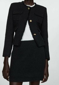Black tweed jacket with gold buttons, features two front pockets, cropped design, and matching black skirt with textured fabric.