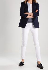 Kvinna står med armarna i kors, iförd en marinblå kavaj, grå- och vitlager på överkroppen, vita byxor och svarta loafers mot en enfärgad bakgrund.