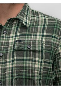 Camisa de franela a cuadros verde y gris con bolsillo en el pecho abotonado y etiqueta de logo. Presenta botones y un cuello extendido. Tejido texturizado.