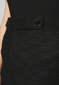 Gros plan sur une jupe noire texturée avec une taille haute ornée d'un bouton rond noir brillant sur le côté.
