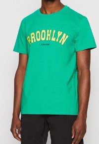 Hombre que lleva una camiseta de manga corta verde con el texto amarillo "BROOKLYN" y el logo negro "KANGOL", combinada con pantalones cortos negros.