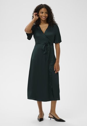 Sötét zöld selyem wrap ruha, rövid ujjakkal, megkötős derékkal és középhosszú szoknyával. Fekete slingback sarkú cipővel viselve.