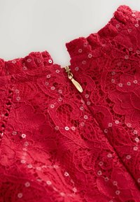 Czerwona koronkowa sukienka z detalami przedstawiającymi teksturowany kwiatowy wzór, udekorowana cekinami oraz złotym zapięciem na plecach.
