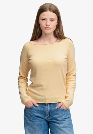 Jonge vrouw met stijlbruin haar draagt een lichtgele lange mouw top en blauwe jeans, staand met handen in de zakken tegen een witte achtergrond.