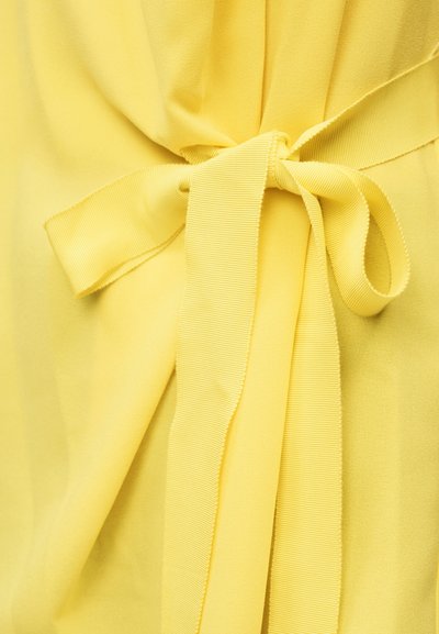 Tissu jaune noué avec un large ruban en gros-grain formant un nœud en forme de boucle, montrant la texture et les plis du matériau.