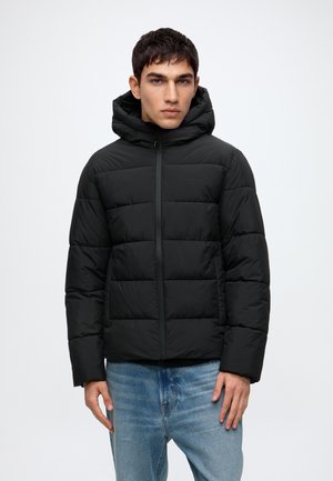 Jeune homme portant une veste matelassée noire à capuche et un jean bleu, debout devant un fond clair uni.
