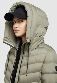Padded jacket in light green with a deep hood, featuring black drawstrings and a zip closure. Accompanied by a black cap.