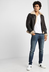 Braune Lederjacke mit einem hellen Fellkragen, kombiniert mit einem weißen T-Shirt und blauen Jeans. Weiße Sneaker mit schwarzen Akzenten runden das Outfit ab.