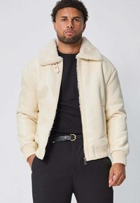 Veste bomber en simili cuir crème avec un col en shearling, des poignets et un ourlet côtelés. Fermeture éclair et poches latérales. Portée par-dessus une chemise noire.