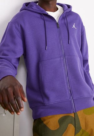 Sudadera morada con cremallera, hecha de suave forro polar. Cuenta con un bolsillo tipo canguro y cordones. Logo en blanco en el pecho, combinada con pantalones de camuflaje.