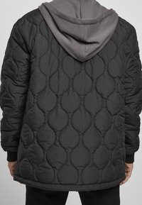 Personne portant une veste matelassée noire avec un sweat à capuche gris en dessous, vue de dos.