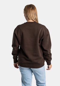 Felpa marrone realizzata in morbido materiale, con una vestibilità rilassata, collo a giro e polsini a costine. Indossata con jeans azzurri chiari.