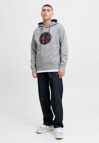 Gray hoodie with a circular black logo and red text, paired with dark jeans and white sneakers with gray accents.
