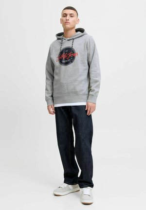 Grijze hoodie met een cirkelvormig zwart logo en rode tekst, gecombineerd met donkere jeans en witte sneakers met grijze accenten.