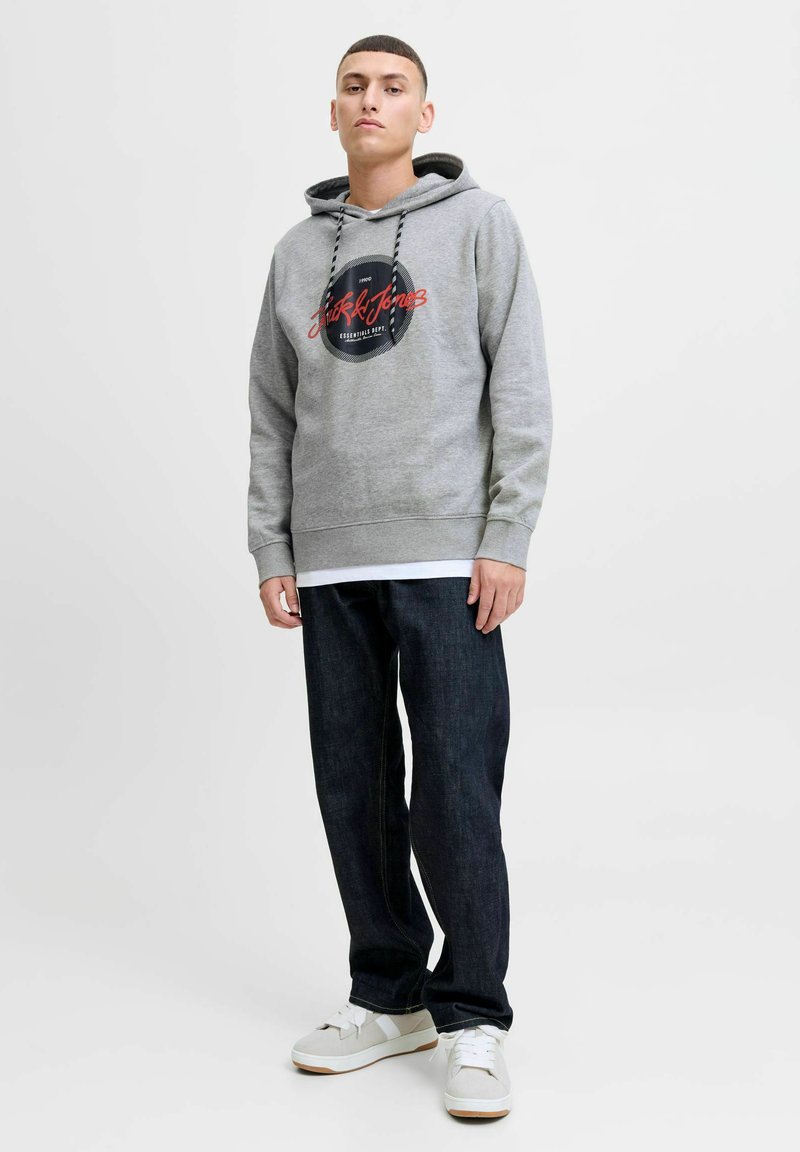 Szary hoodie z okrągłym czarnym logo i czerwonym napisem, w zestawieniu z ciemnymi dżinsami i białymi trampkami z szarymi akcentami.