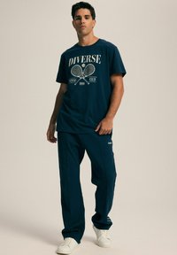 Granatowa bawełniana koszulka z nadrukiem "Diverse Tennis Club 1998", zestawiona z pasującymi granatowymi spodniami. Białe sneakersy dopełniają stylizację.