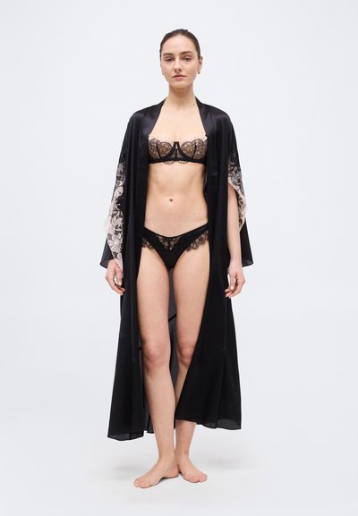 Γυναίκα όρθια ξυπόλητη, φορώντας μαύρα δαντελένια εσώρουχα και μακρύ μαύρο σατέν ρόμπα με δαντελένια μανίκια, μπροστά σε απλό λευκό φόντο.