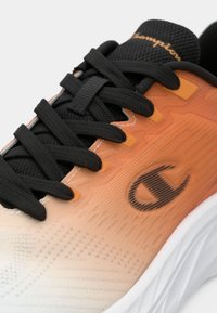 Πορτοκαλί και λευκό αθλητικό παπούτσι με υφή στην επιφάνεια, μαύρα κορδόνια και μαύρη ταινία έλξης, με λογότυπο στο πλάι.