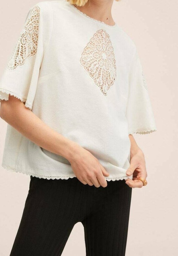 Femme portant un haut blanc à manches courtes avec des motifs en dentelle ajourée sur les manches et la poitrine, associé à un pantalon côtelé noir.