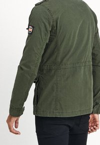 Olivgrön kanvasjacka med en strukturerad design, som har ett märke på vänster axel och elastiska ärmslut. Enkel och funktionell stil.