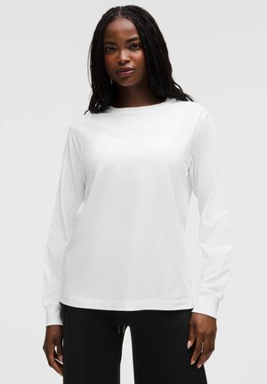 Femme aux longs cheveux tressés portant une chemise blanche unie à manches longues et un pantalon noir, se tenant devant un fond gris clair.