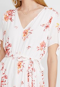 Vit blommig klänning med rosa och orange mönster, korta ärmar, en djup V-ringning och ett tygbälte i midjan. Mjuk textur.