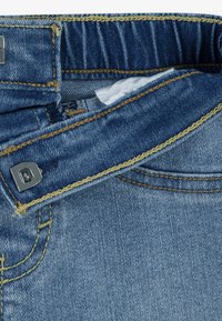 Κοντινό πλάνο τζιν με μπλε απόχρωση, που διαθέτει αντίθετη κίτρινη ραφή, ασημένια εξαρτήματα και απαλό εσωτερικό επένδυση.