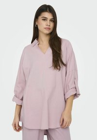 Chemise ample de couleur rose clair avec un col en V, des manches repliées et une texture douce ; fabriquée à partir d'un tissu léger adapté pour le superposition.
